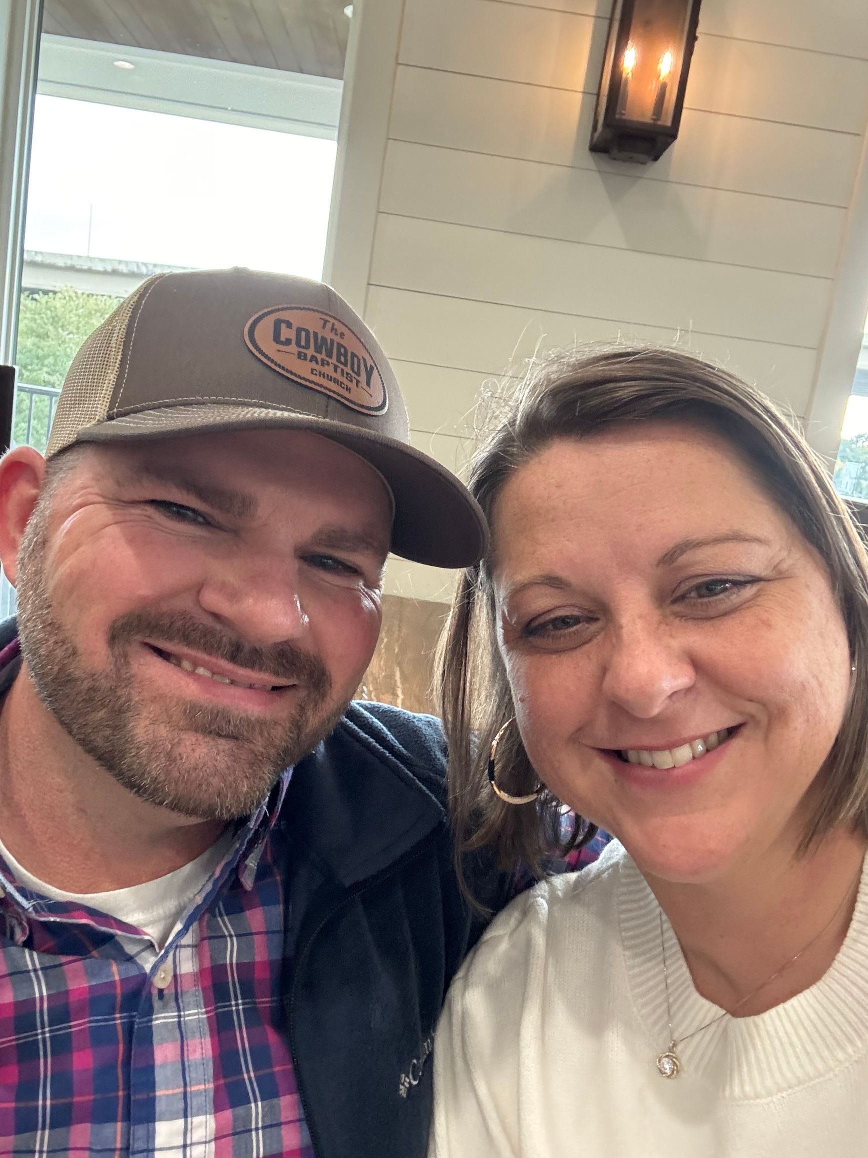 Smiling couple, man in plaid shirt and cap, woman in dark shirt, indoors.