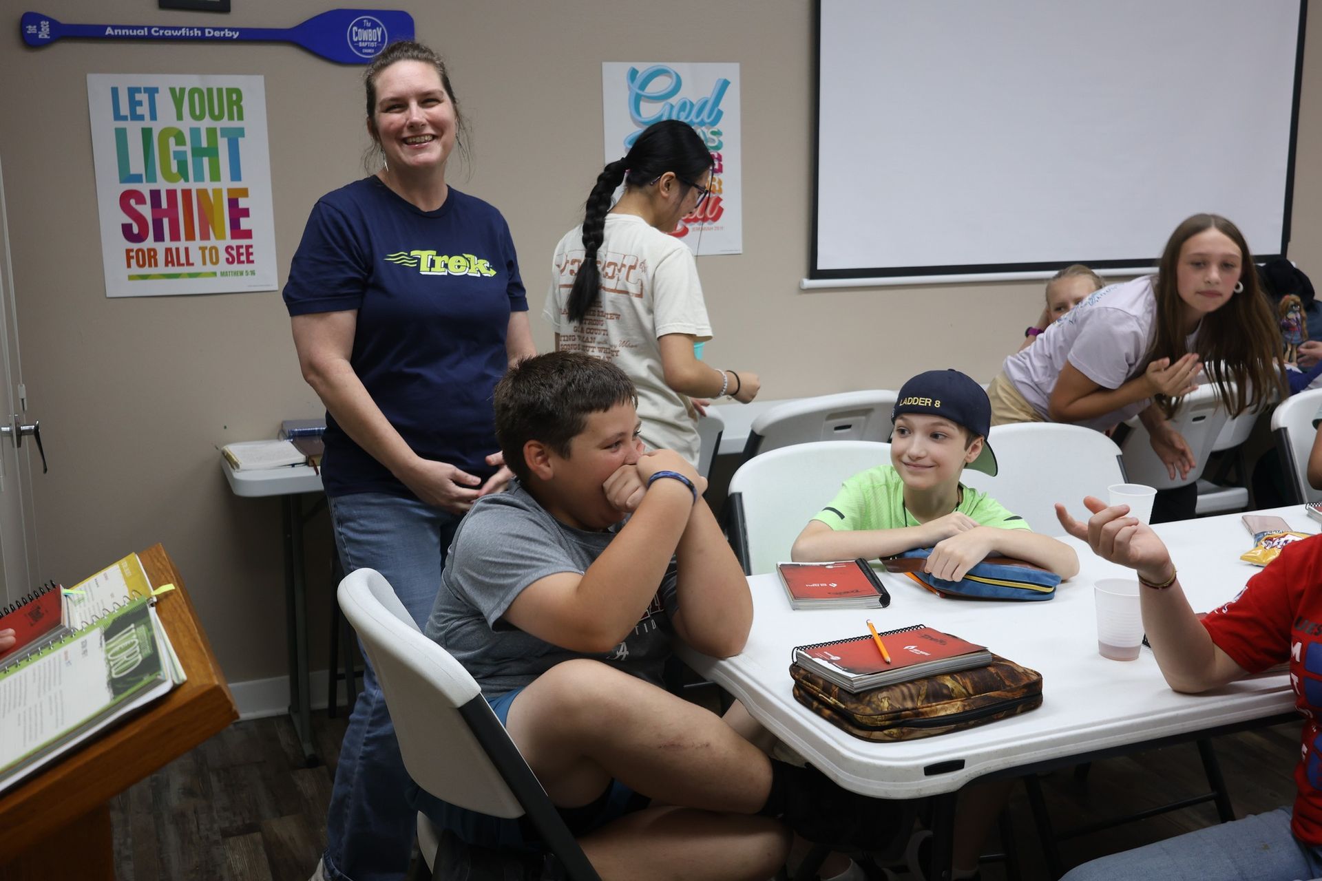 People at a table with cake. Woman in blue shirt smiles, others watch. Room with projector screen, posters.