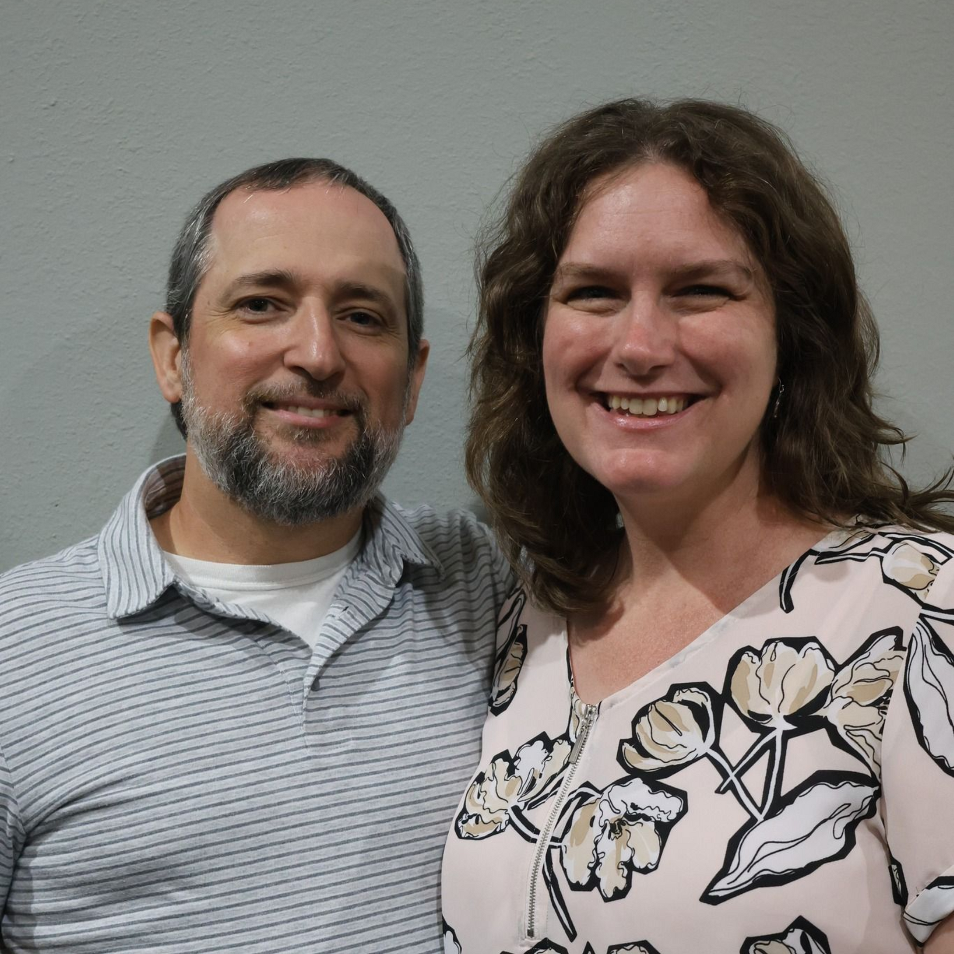 Man and woman smiling, posed together. Man wears striped shirt, woman floral top. Against plain wall.