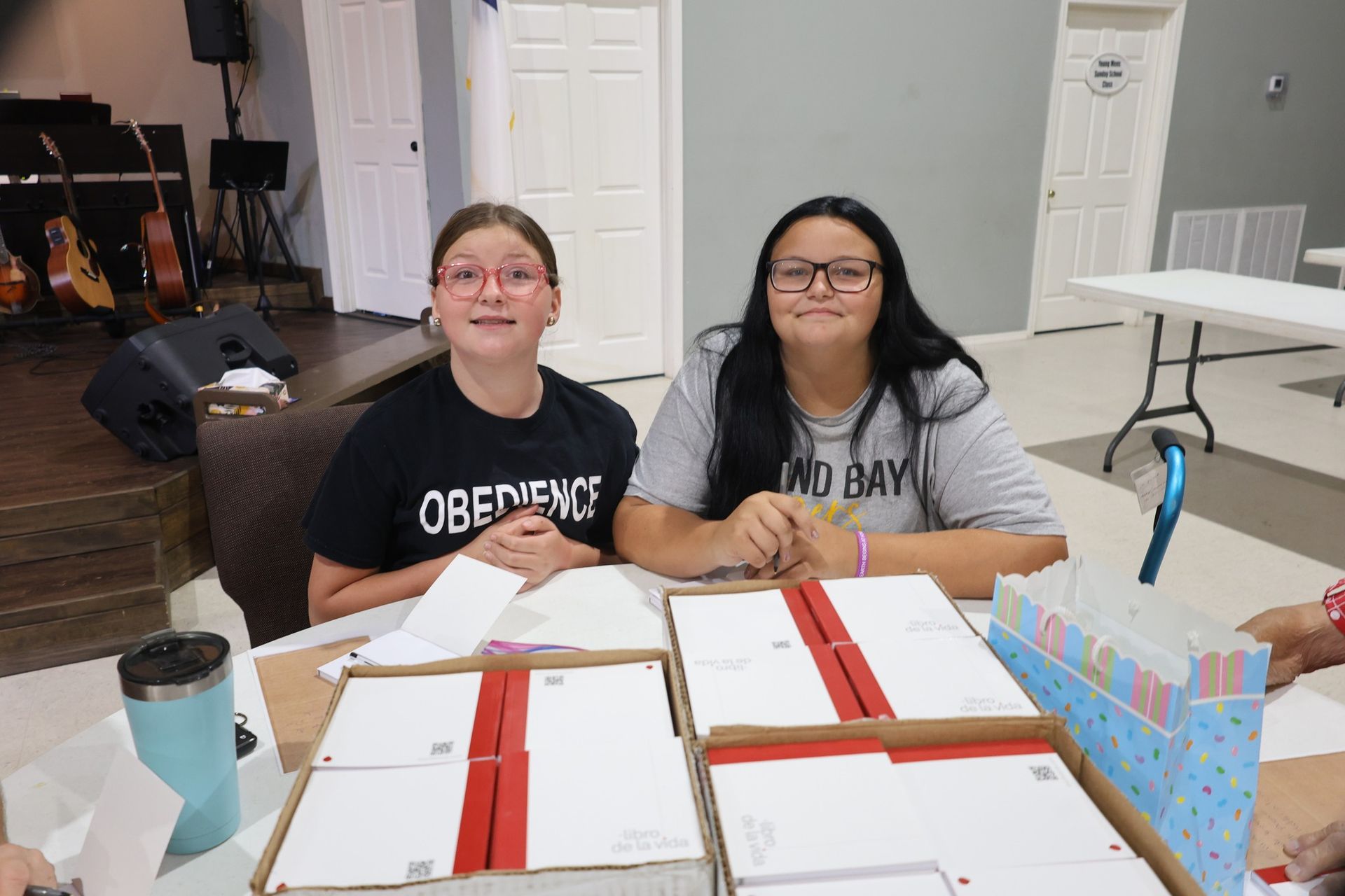 Two people seated at a table, packaging boxes. Inside a building with a stage and instruments.