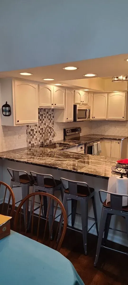 A kitchen with white cabinets , granite counter tops , stools and a table.
