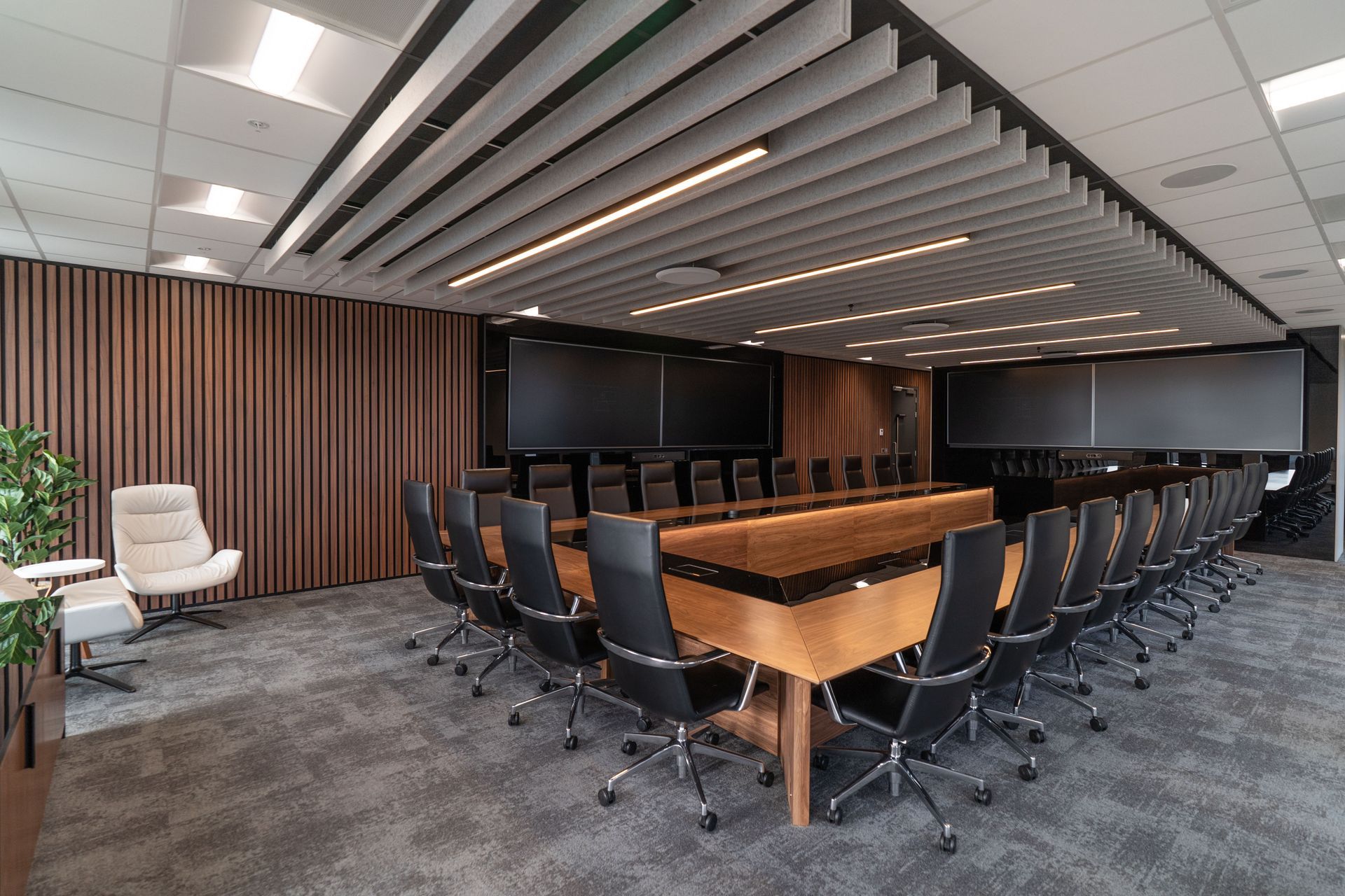 Conference room with a large wooden table, chairs, and multiple screens. Dark wood panelling and grey carpet.