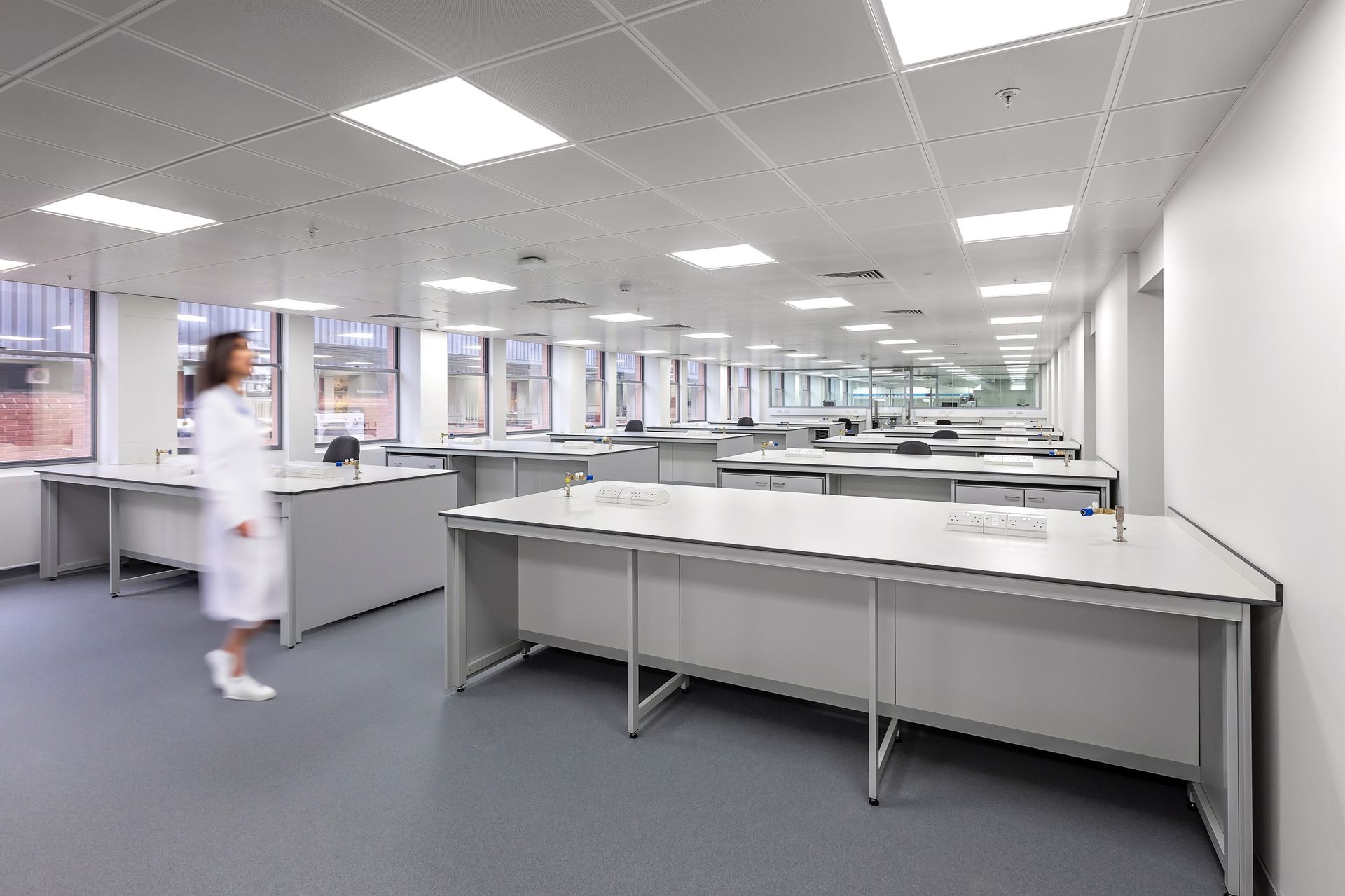 A woman in a lab coat is walking through a large room filled with desks.