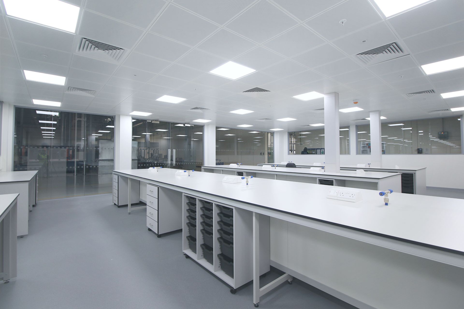 Bright laboratory with white countertops, cabinets, and ceiling lights, a grey floor, and glass walls.