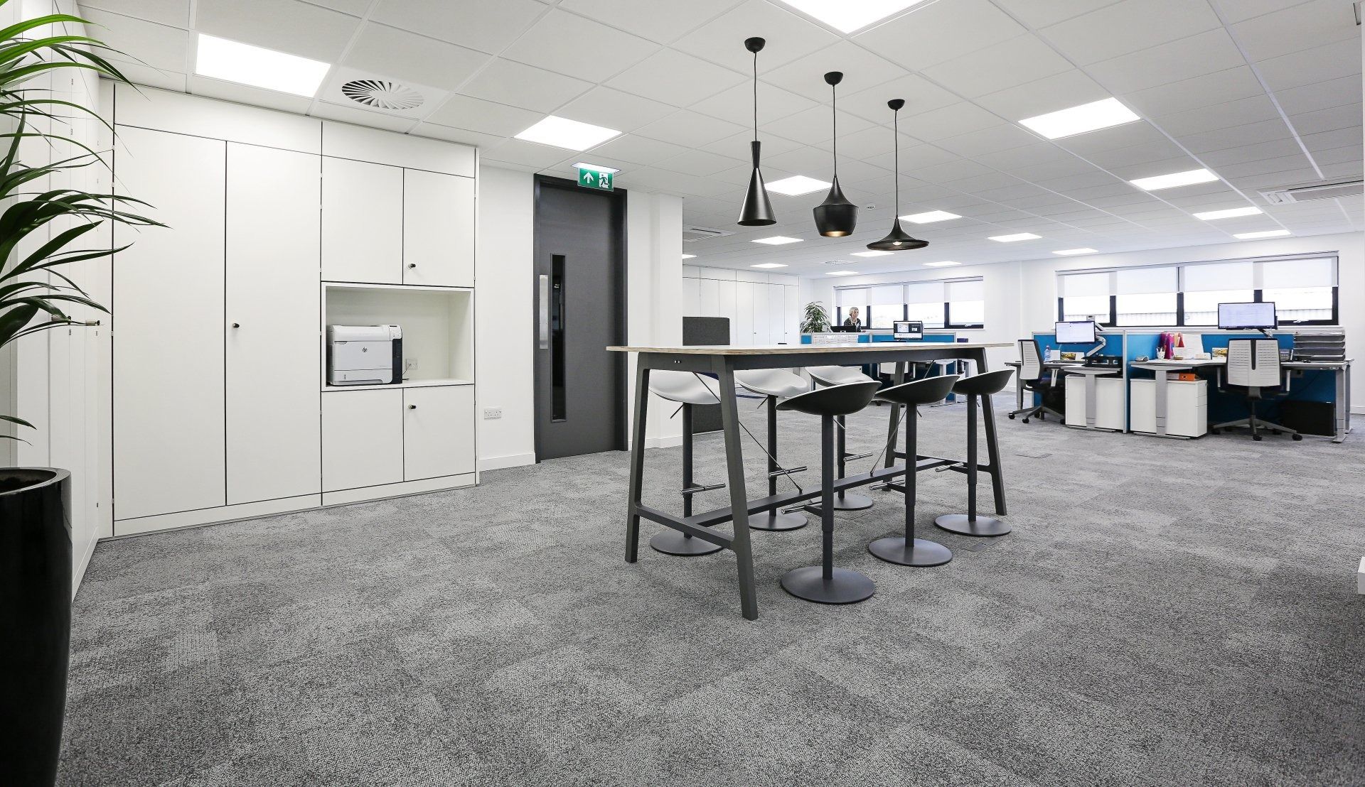 Bright office space with a high table, stools, and pendant lights. White cabinets and cubicles are visible.