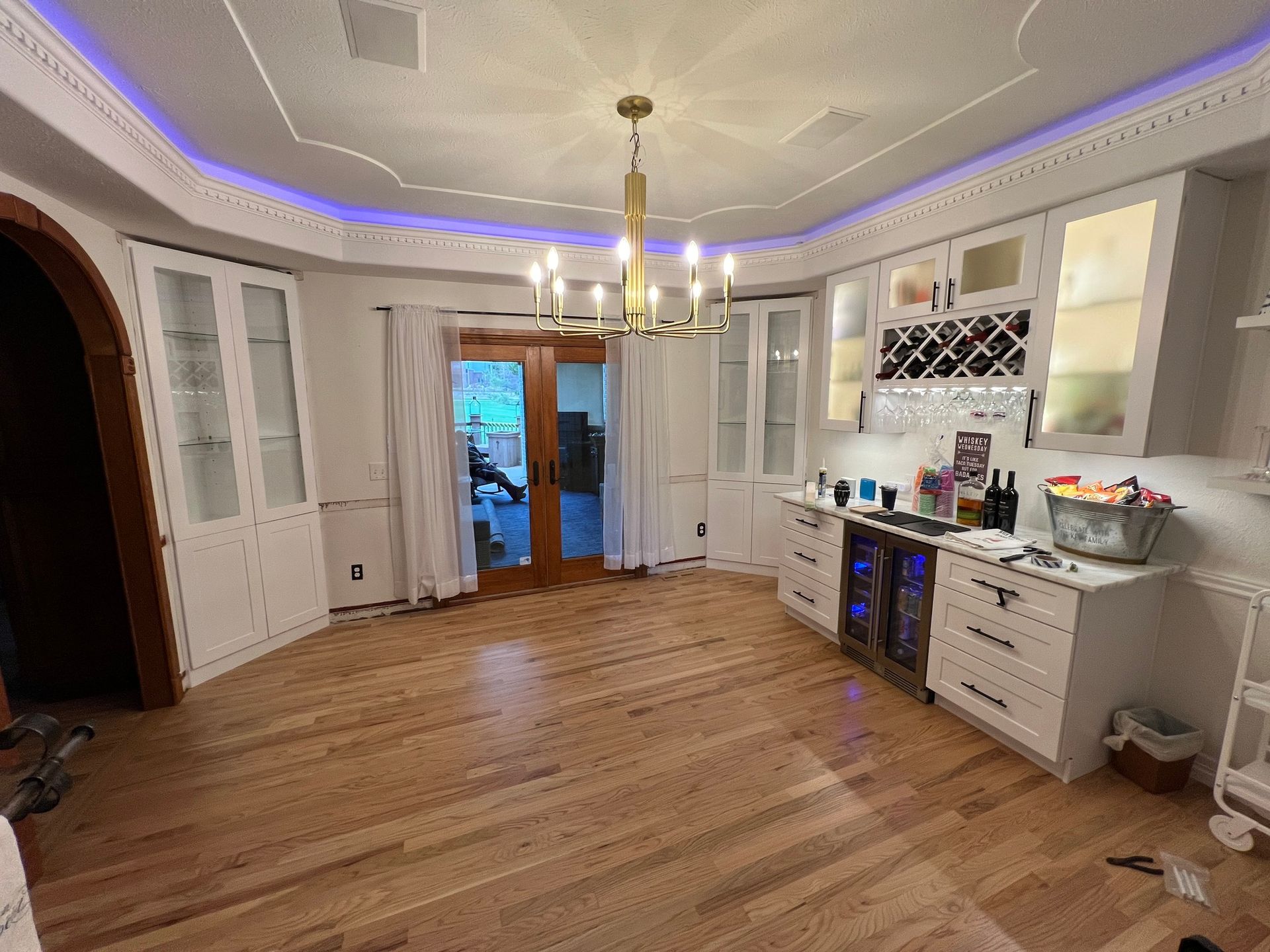 A dining room and wet bar area with white cabinetry, wooden floors, a gold chandelier, and blue crown molding lighting.