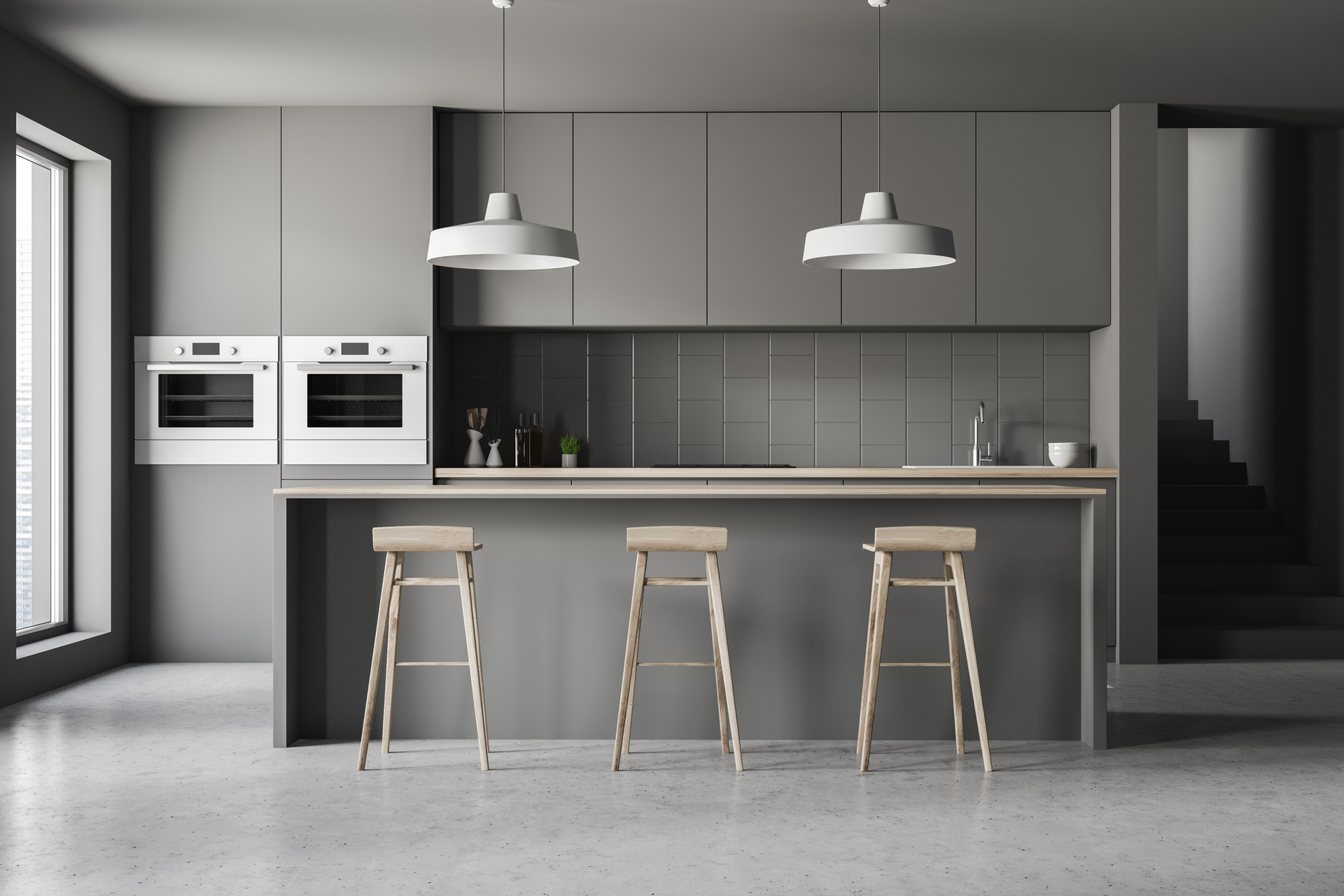 Modern gray kitchen with a breakfast bar, three wooden stools, white wall ovens, and two minimalist pendant lights.