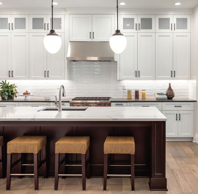 A kitchen with a dark brown island, woven stools, white cabinets, stainless appliances, and a marble-topped counter.