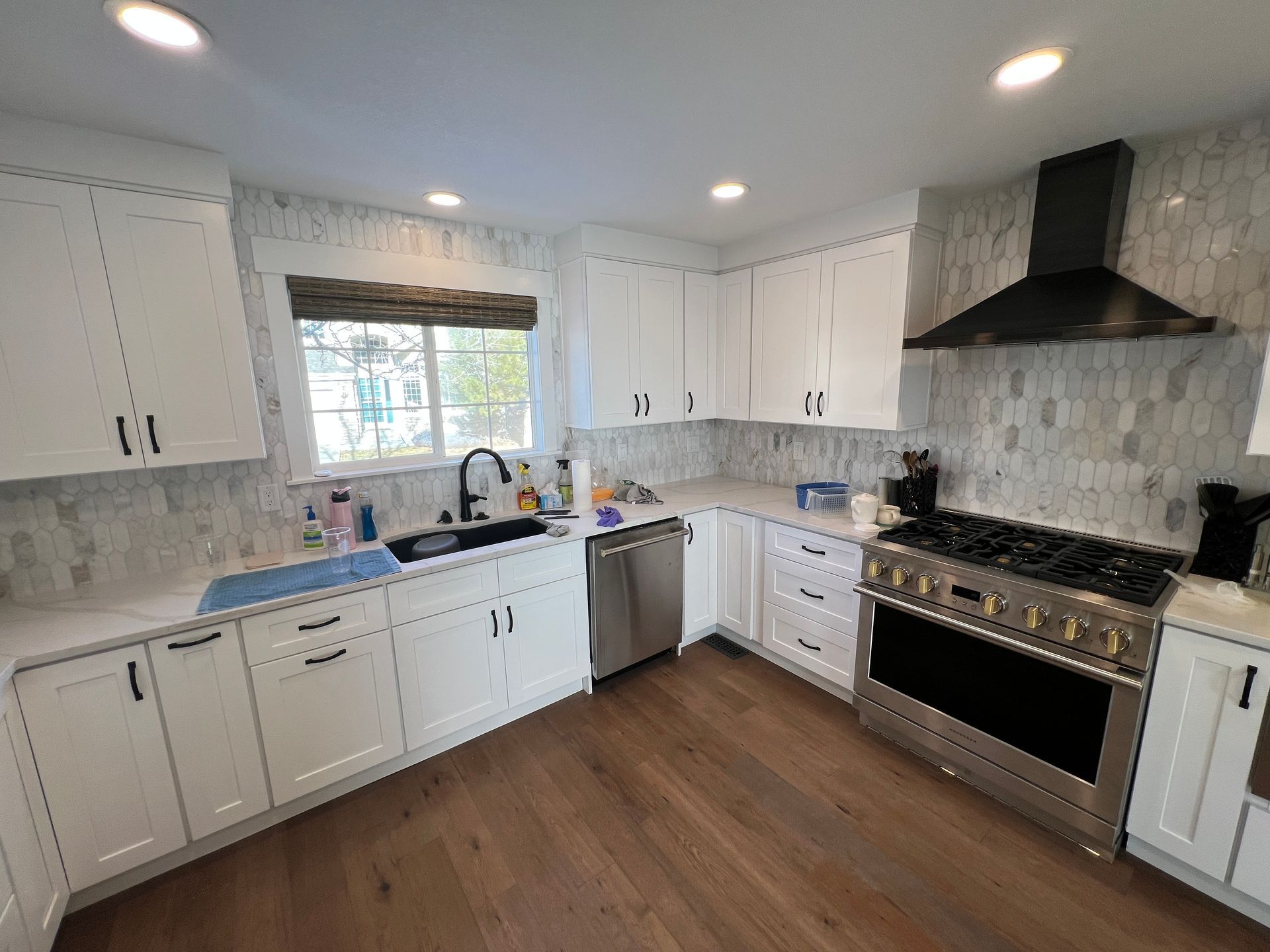 A modern kitchen with white cabinets, a large stainless steel gas range with a black hood, and light wood flooring.