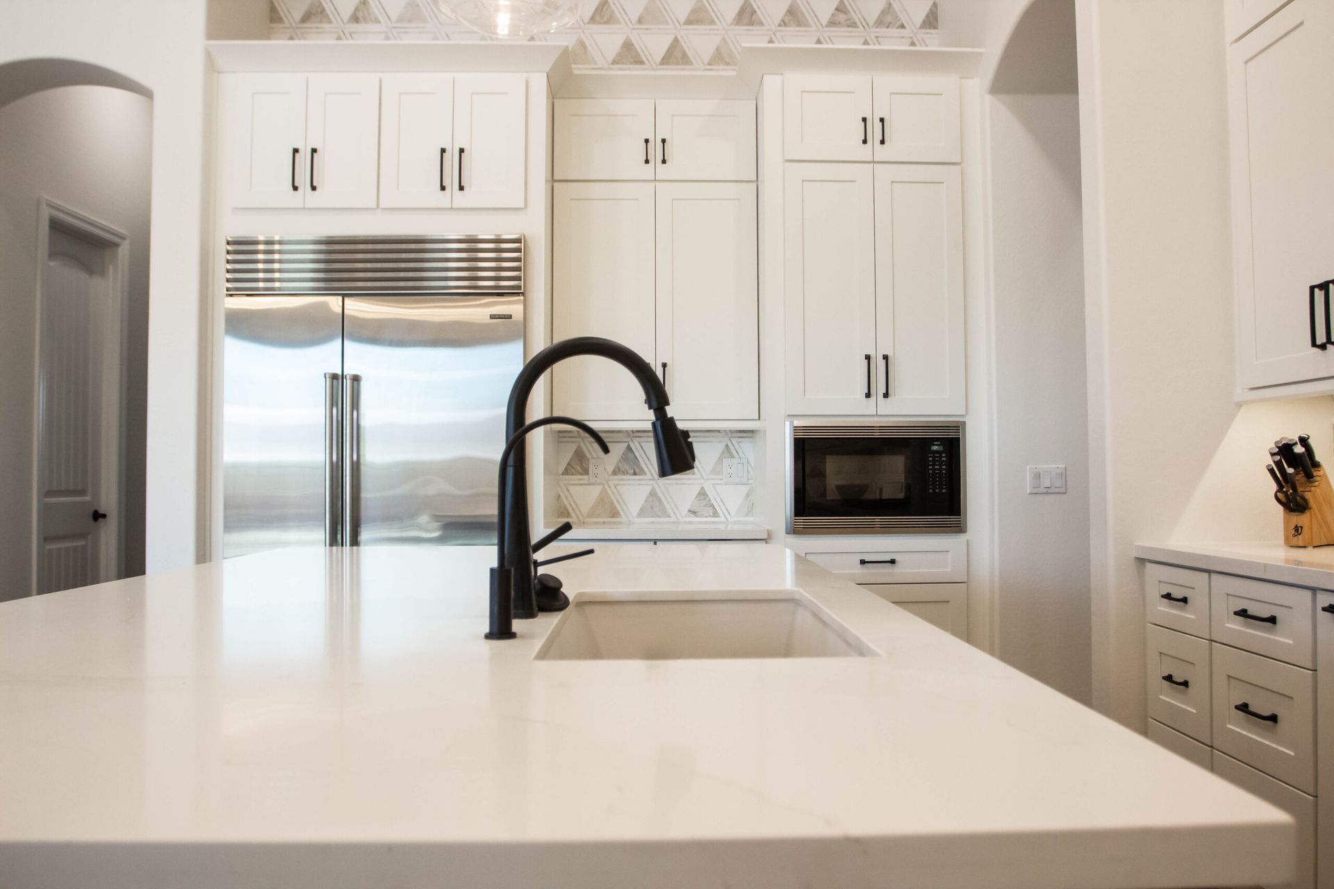 A modern kitchen with white cabinets, a large white quartz island, a matte black faucet, and a built-in microwave.