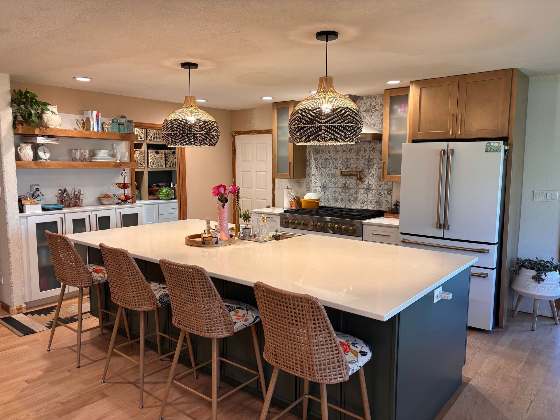 A kitchen with a large white island, four wicker bar stools, two pendant lights, and light wood cabinets.
