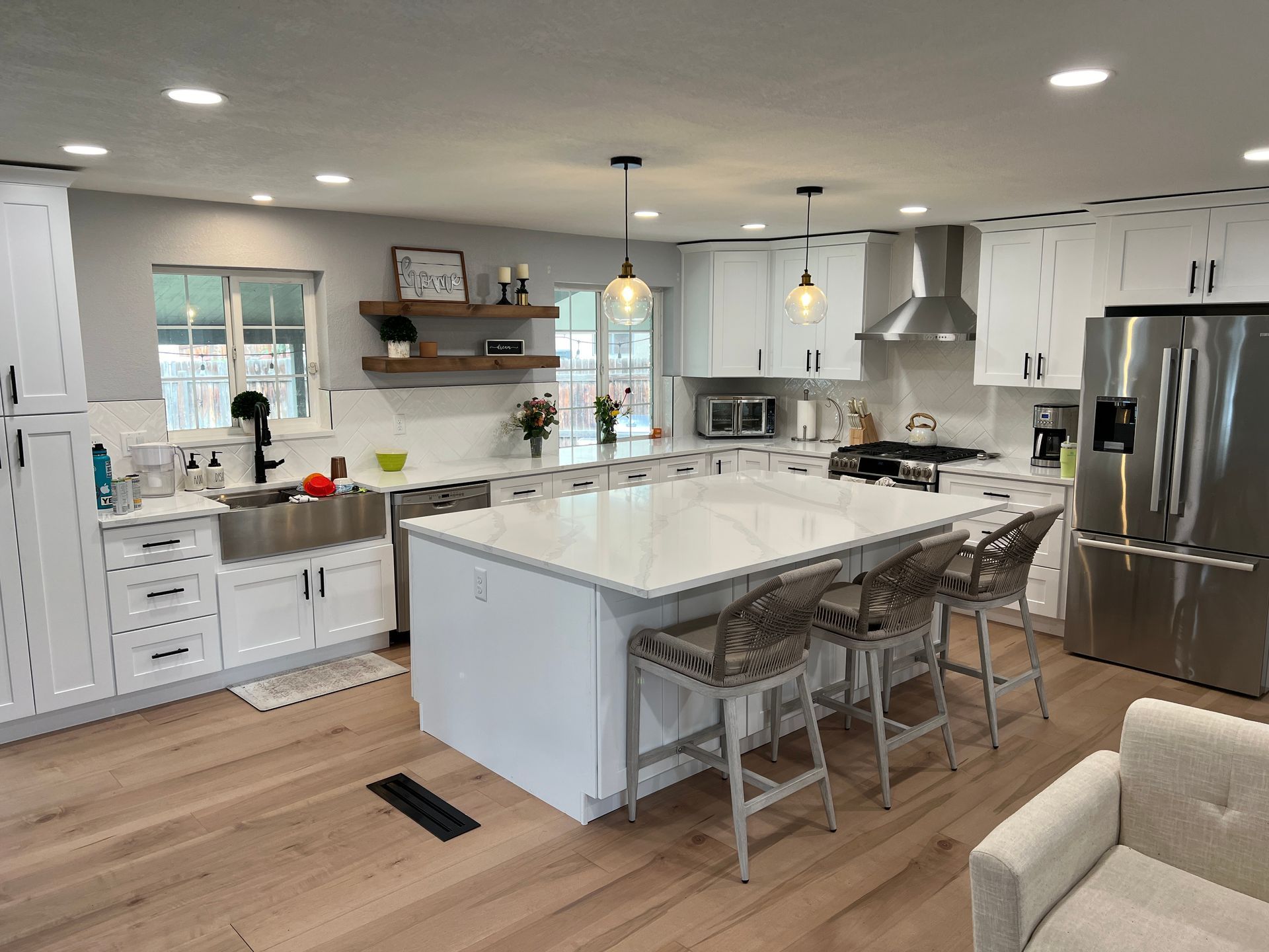 A modern white kitchen featuring a large marble-topped island with three bar stools and light wood flooring.