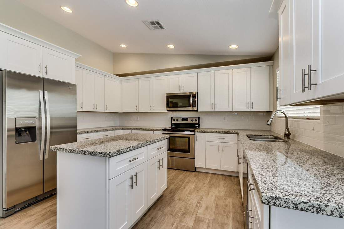 A bright, modern kitchen featuring white cabinets, speckled granite countertops, stainless steel appliances, and wood floors.