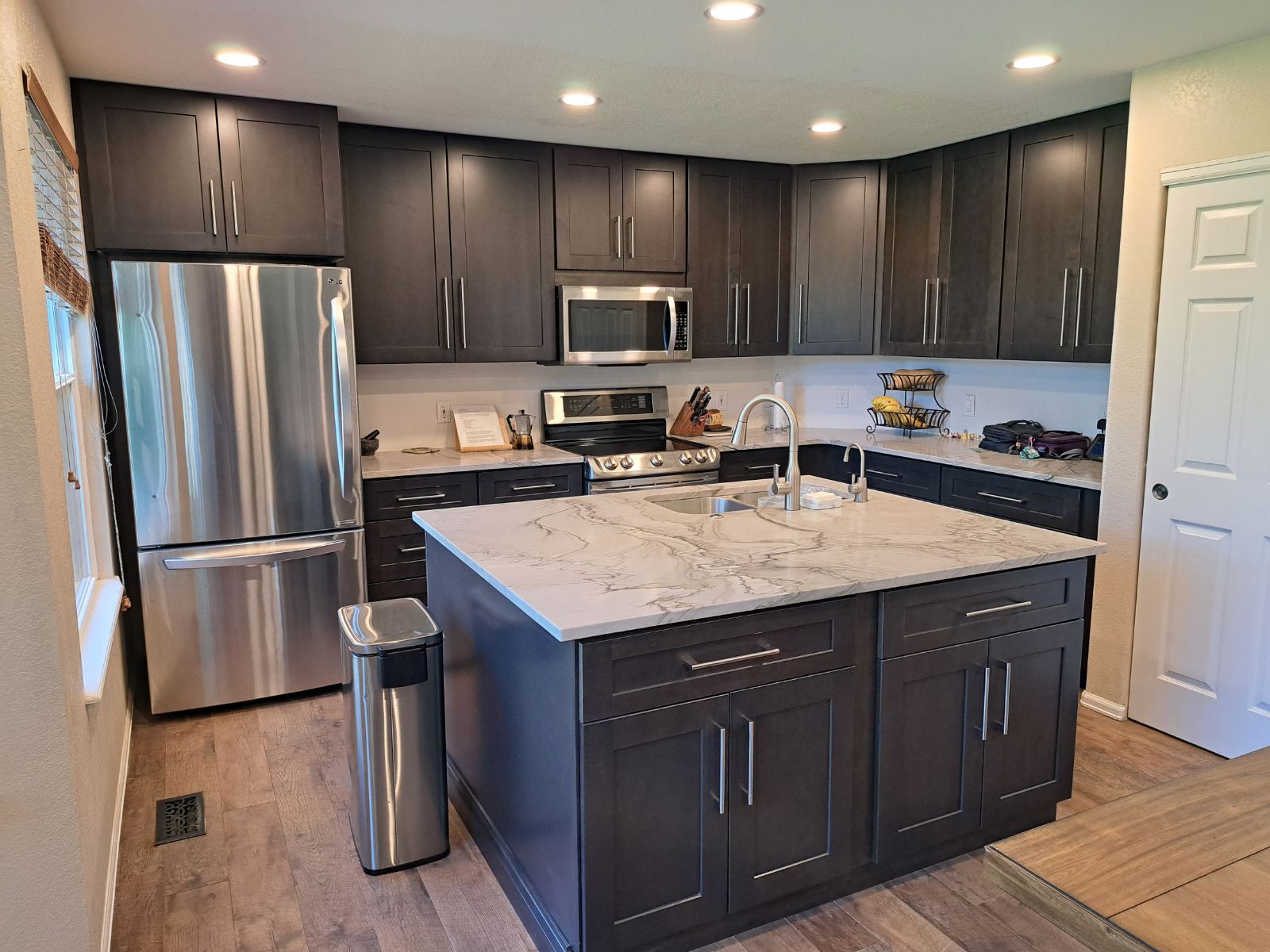A kitchen with dark wood cabinets, stainless steel appliances, a marble-topped island, and light wood flooring.