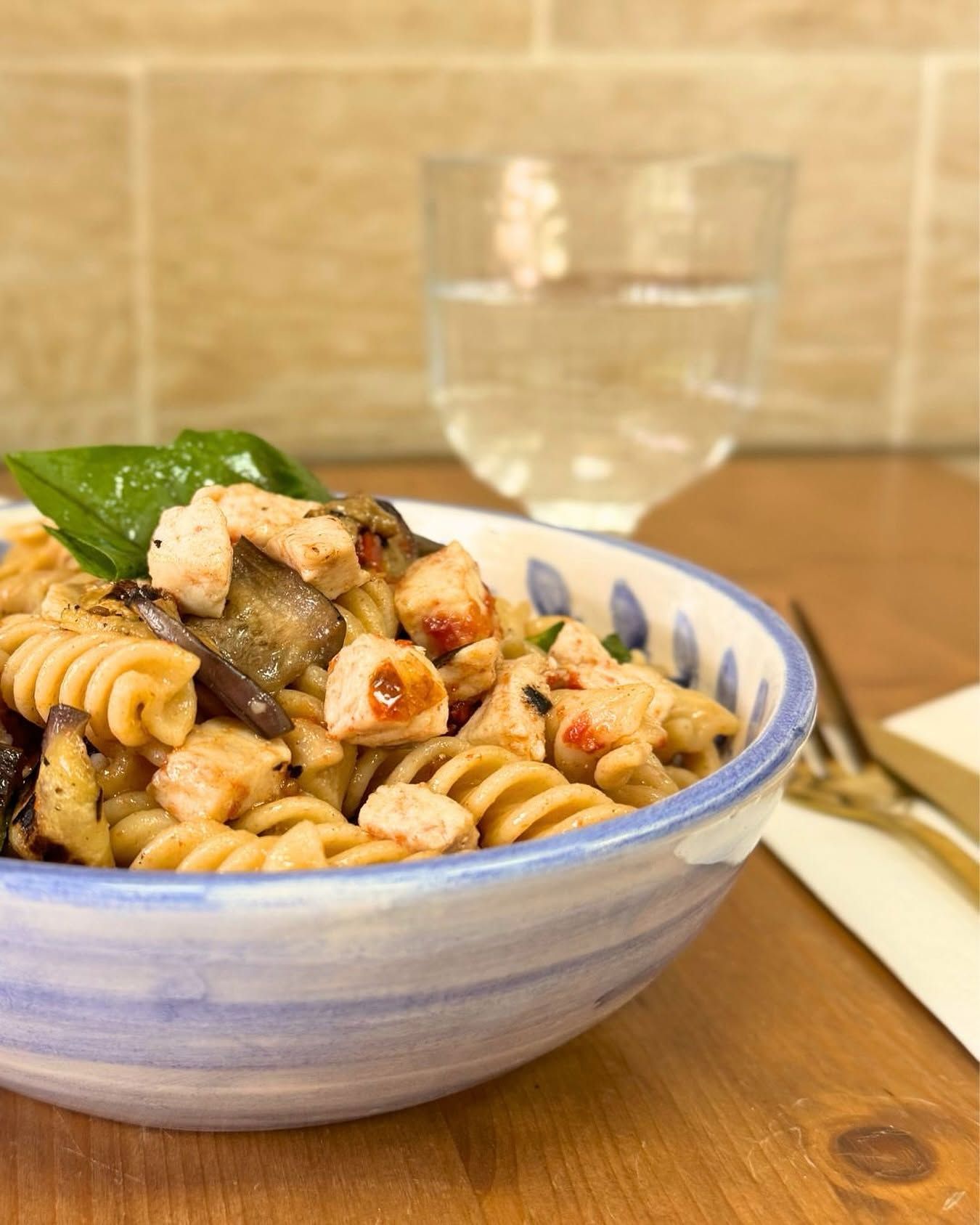 Una ciotola di pasta rotini con pollo e basilico su un tavolo di legno, con un bicchiere d'acqua e una forchetta nelle vicinanze.