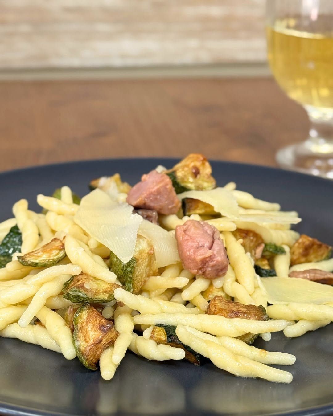Piatto di pasta al pesto, carne scottata e formaggio grattugiato su un piatto scuro, con una bevanda sullo sfondo.