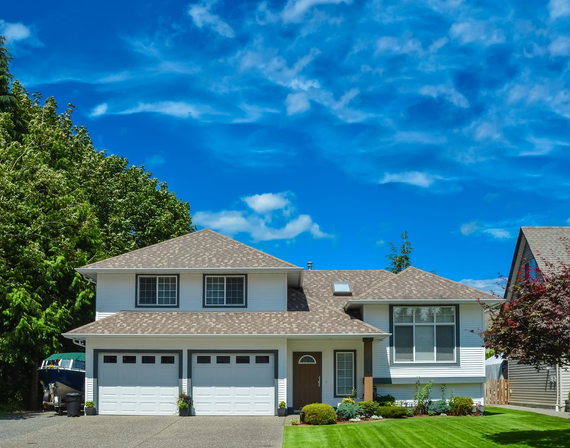 A two-story white house with a dark shingled roof, two garage doors, and a green lawn under a bright blue sky.