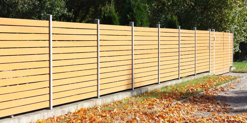 A horizontal wooden slat privacy fence with grey metal posts sitting on a concrete base, bordered by fallen autumn leaves.
