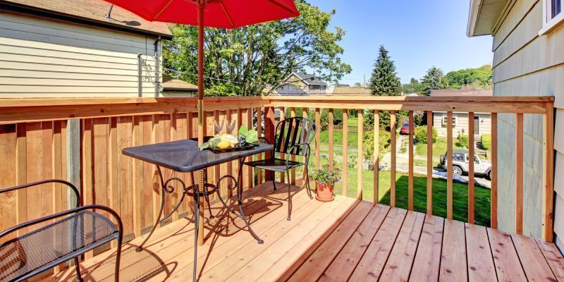 A wooden deck featuring a small table with a red umbrella, two metal chairs, and a potted plant, overlooking a yard.