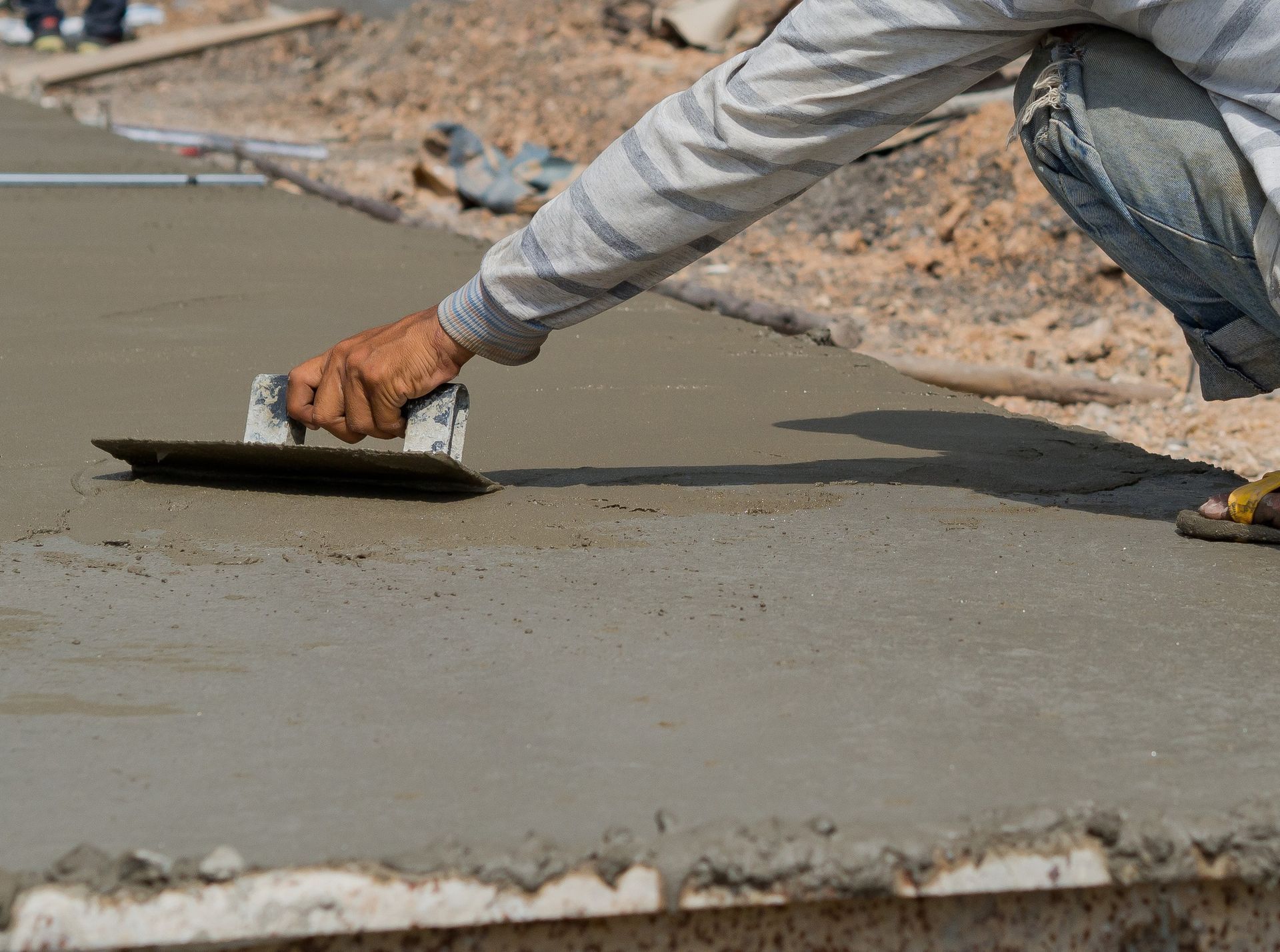 A construction worker crouches while using a trowel to smooth wet, grey concrete on a job site.