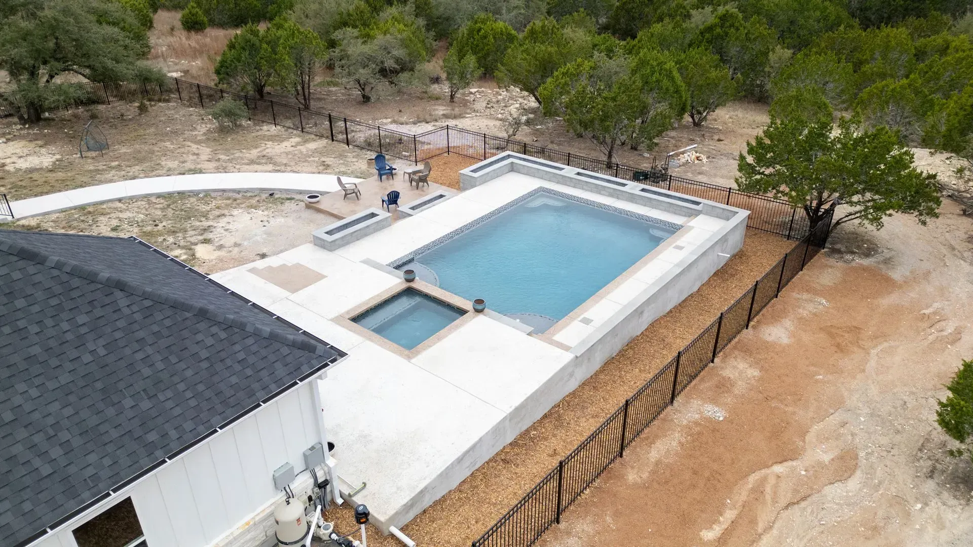 An aerial view of a rectangular swimming pool and attached hot tub on a concrete patio next to a house in a wooded area.