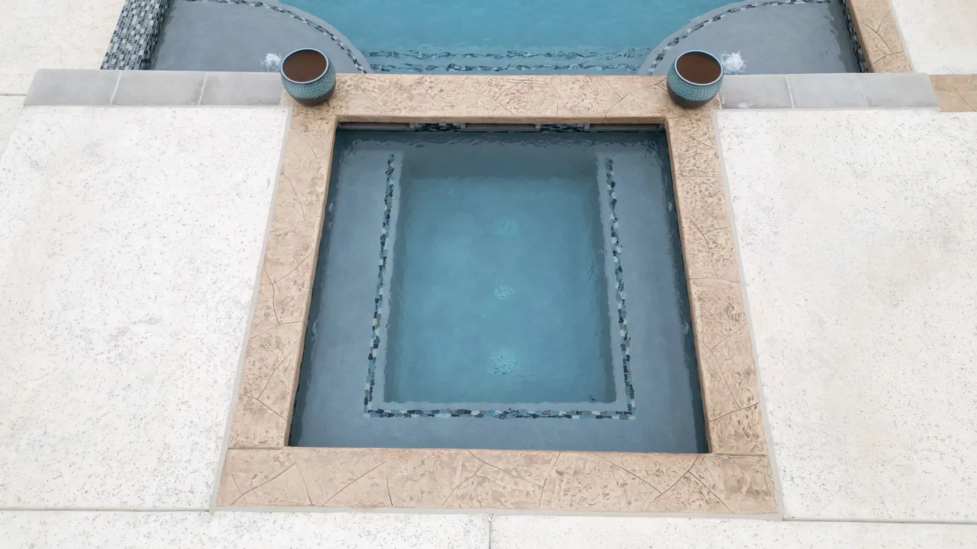 A rectangular spa with blue water and stone tiling, set into a beige stone patio adjacent to a swimming pool.