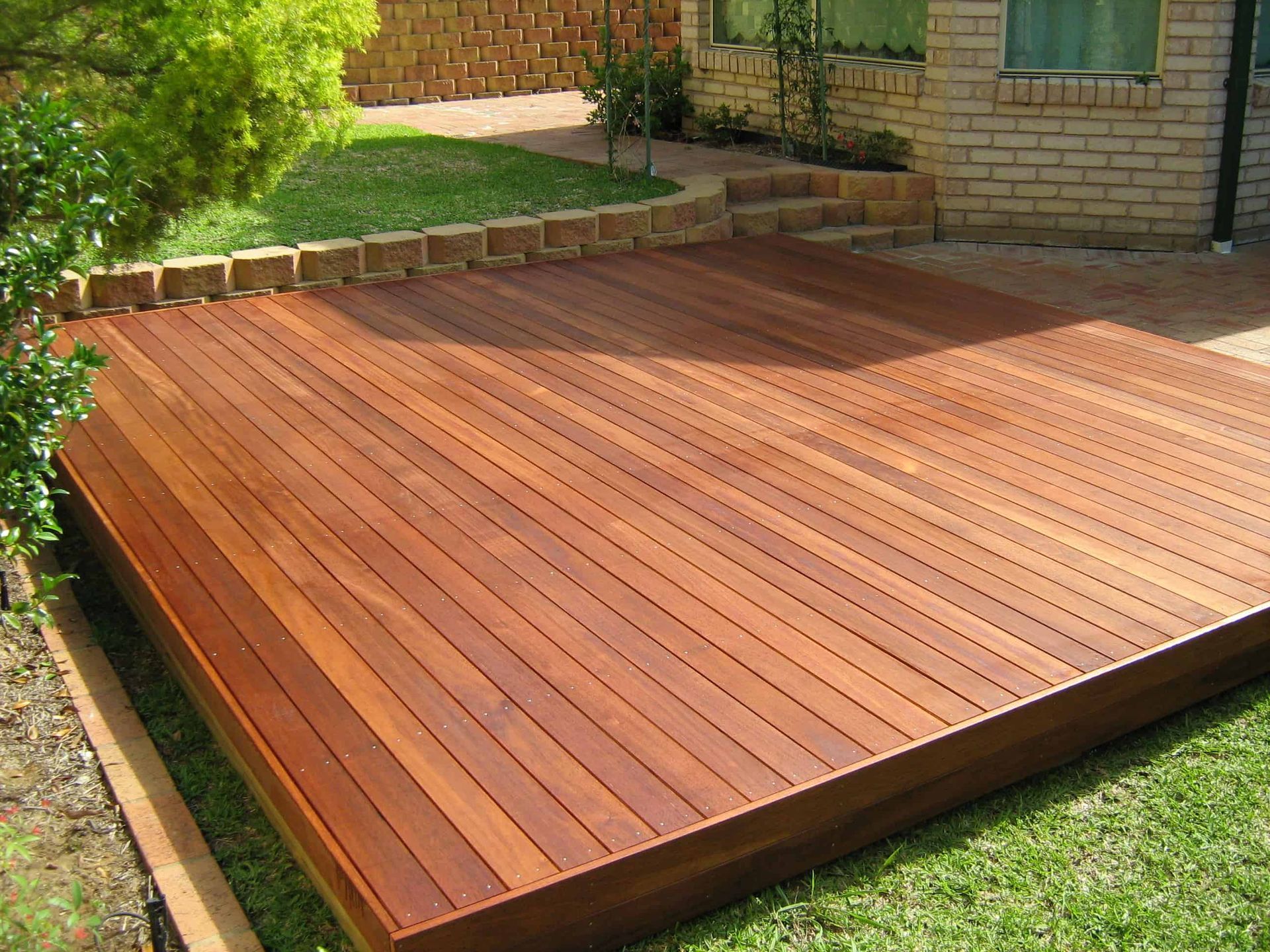 A rectangular wooden deck made of reddish-brown planks sits in a backyard next to a brick house and grass.