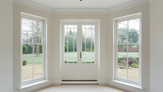A set of white double doors flanked by two tall sash windows in a room with cream walls and a view of a garden.