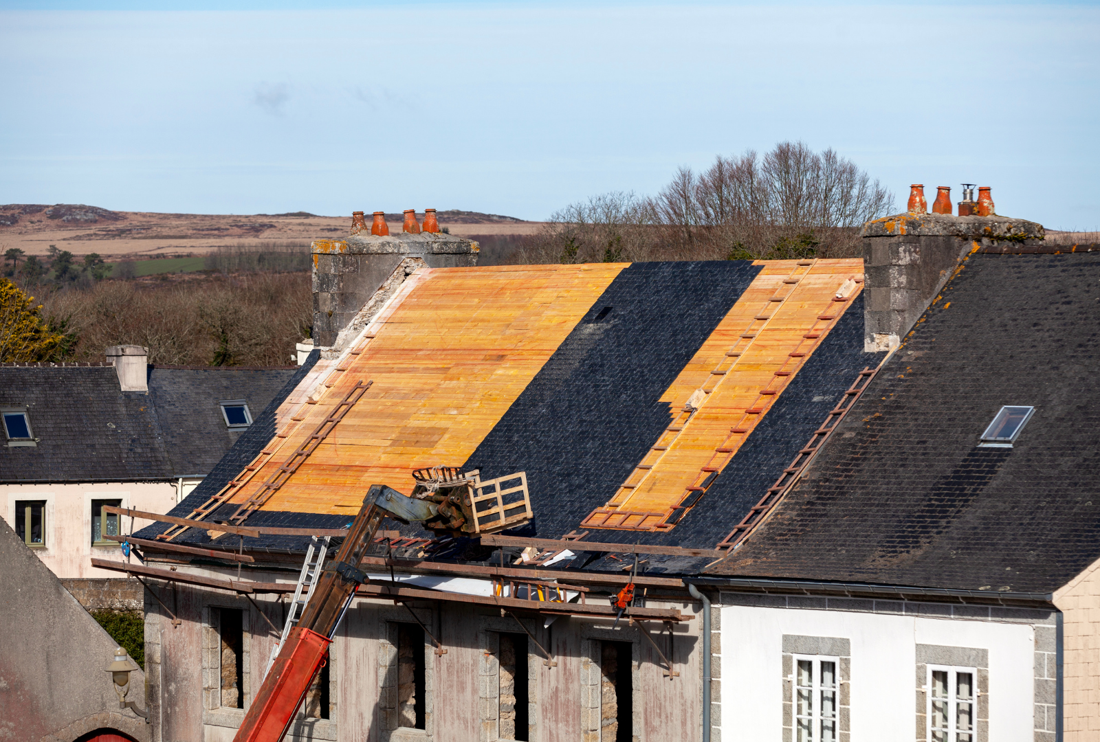 A partially stripped roof with wooden decking and black underlay, undergoing repairs, with a crane arm visible.