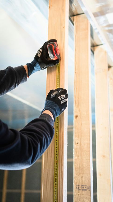A person wearing work gloves measures a wooden wall stud with a yellow tape measure during construction.