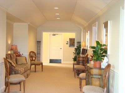 Hallway with beige walls, carpet, and ornate furniture; potted plants and a closed door are visible.