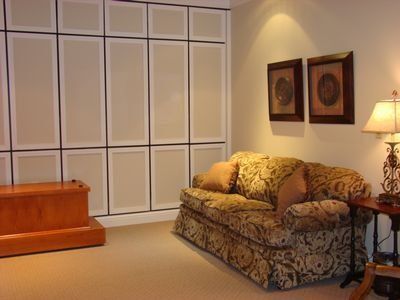 Beige patterned couch and artwork in a room with a large paneled wall and a wooden chest.