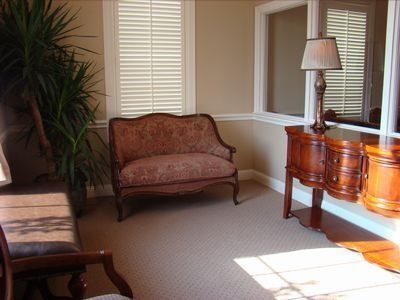 Cozy room with loveseat, end table, lamp, and plant. Natural light streams through window shutters.