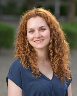 A woman with red curly hair is wearing a blue shirt and smiling for the camera.