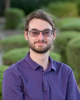 A young man with a beard and glasses is wearing a purple shirt.