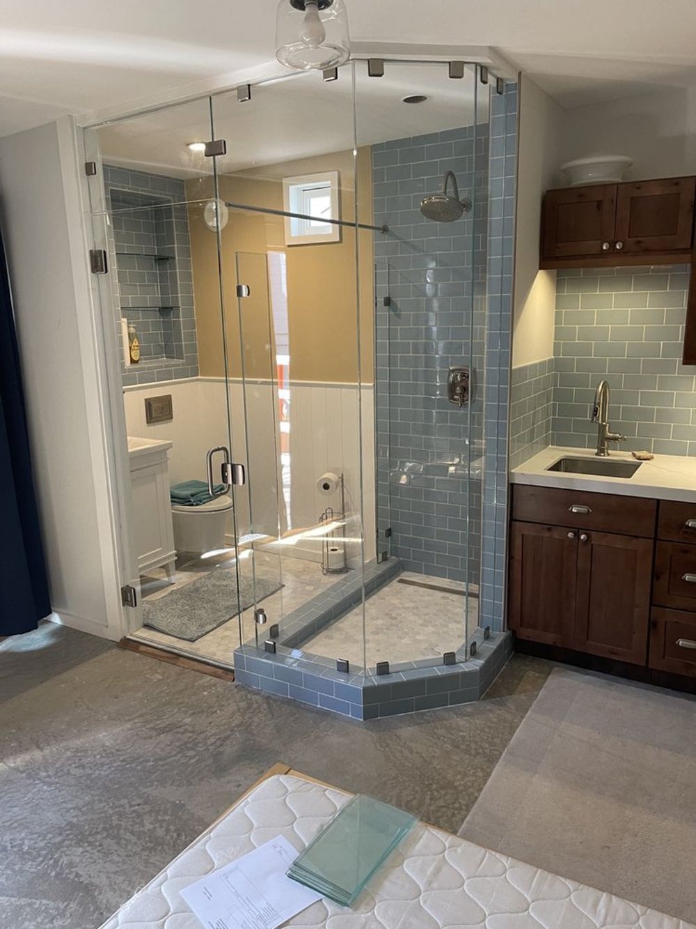 Shower stall with glass walls next to a small kitchenette. The stall is blue-tiled, the cabinets are dark brown.
