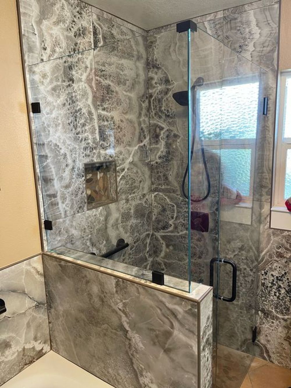Gray marble-tiled shower with glass door and window. Features black hardware and a built-in shelf.