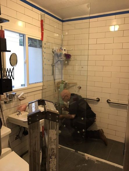 Man kneeling in a shower stall fixing something. White tiled walls, safety bars, window.