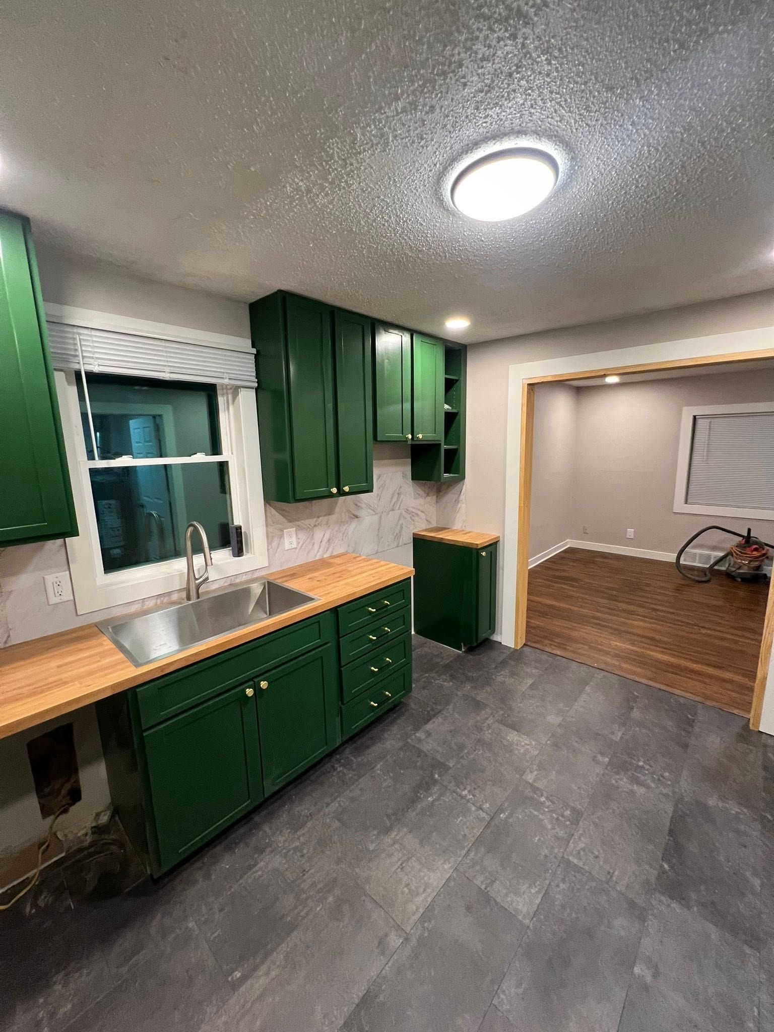 Green kitchen cabinets with wooden countertops, slate floor, and a view into a room with wood flooring.