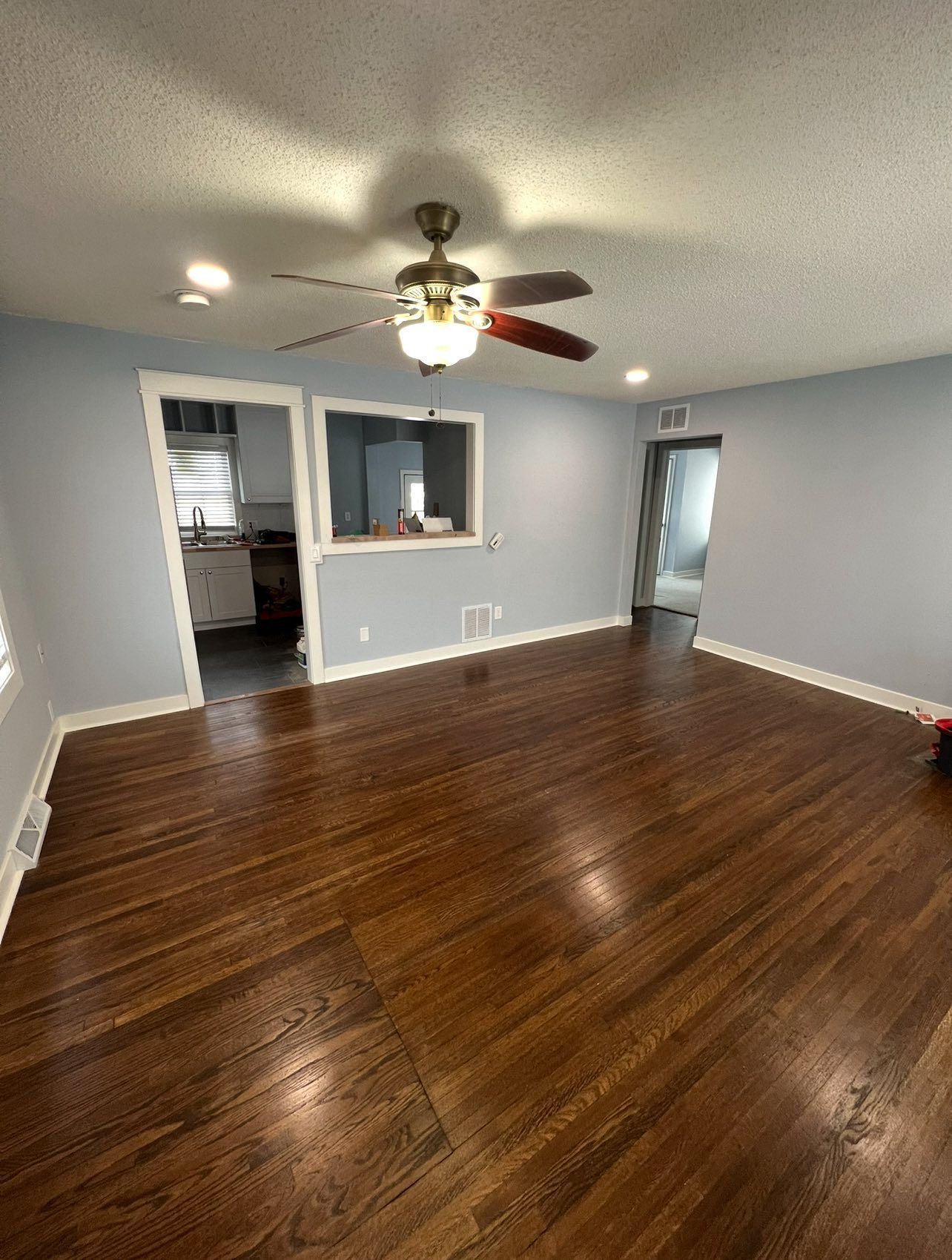 Living room with hardwood floor, light blue walls, and a ceiling fan. Kitchen visible through an opening.