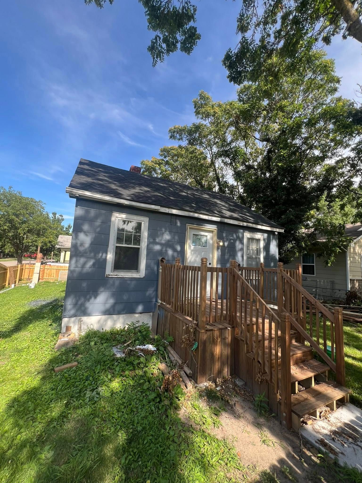 Small, weathered blue house with wooden porch and stairs, surrounded by green grass and trees under a blue sky.