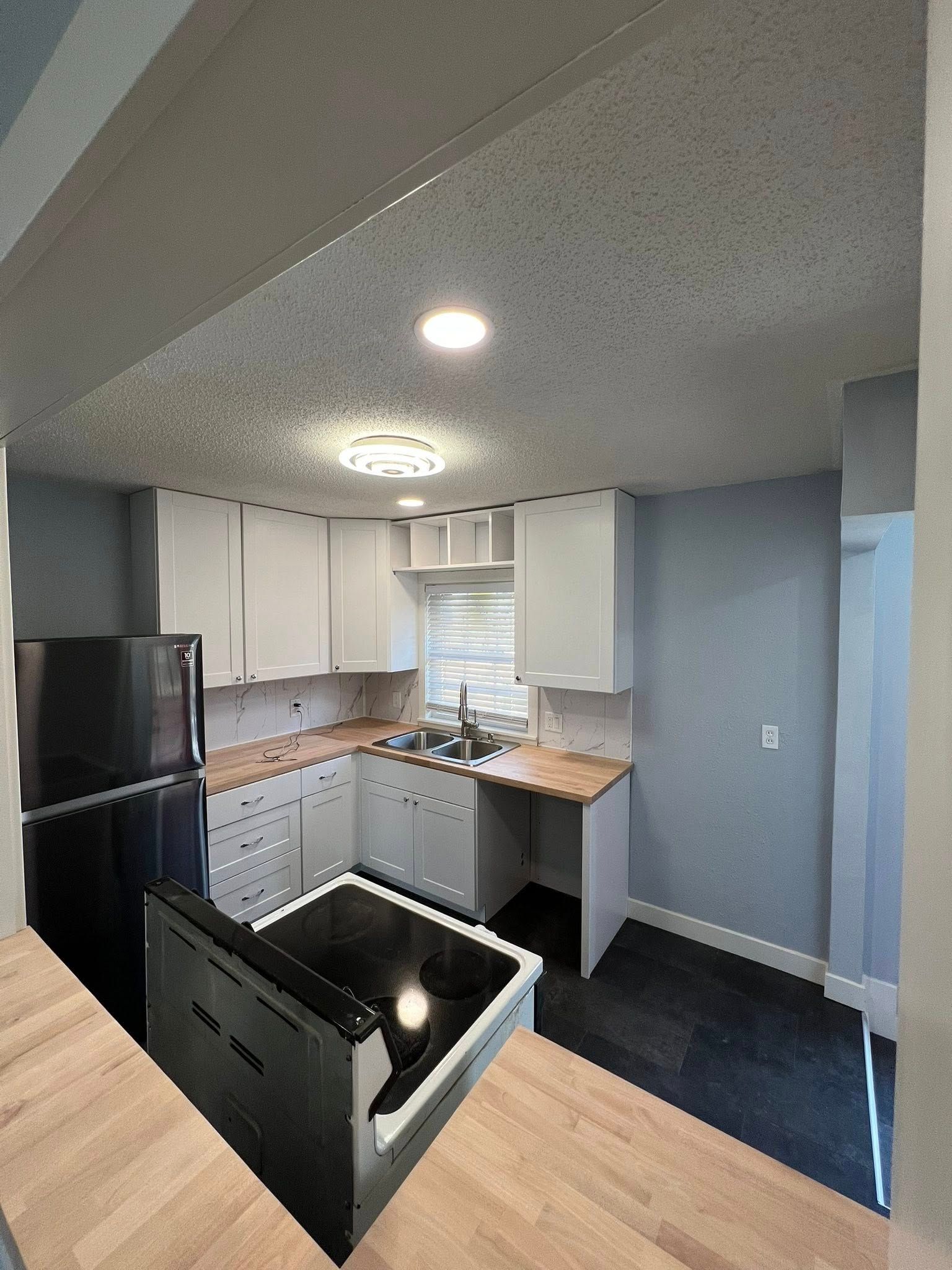 Small, modern kitchen with white cabinets, light countertops, and black appliances. Dark floor, light walls.