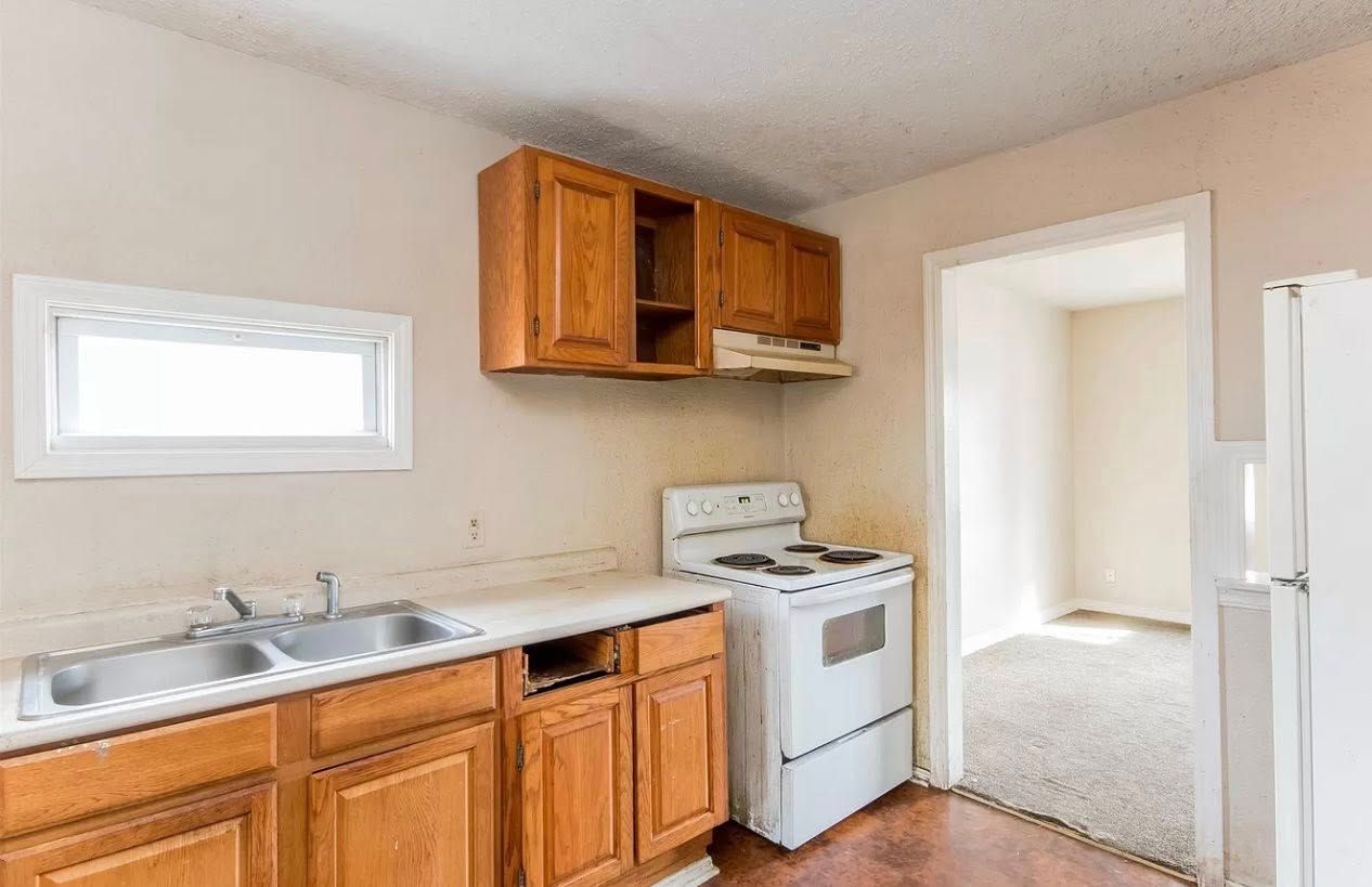Kitchen with wooden cabinets, white appliances, and an open doorway to another room with carpet.