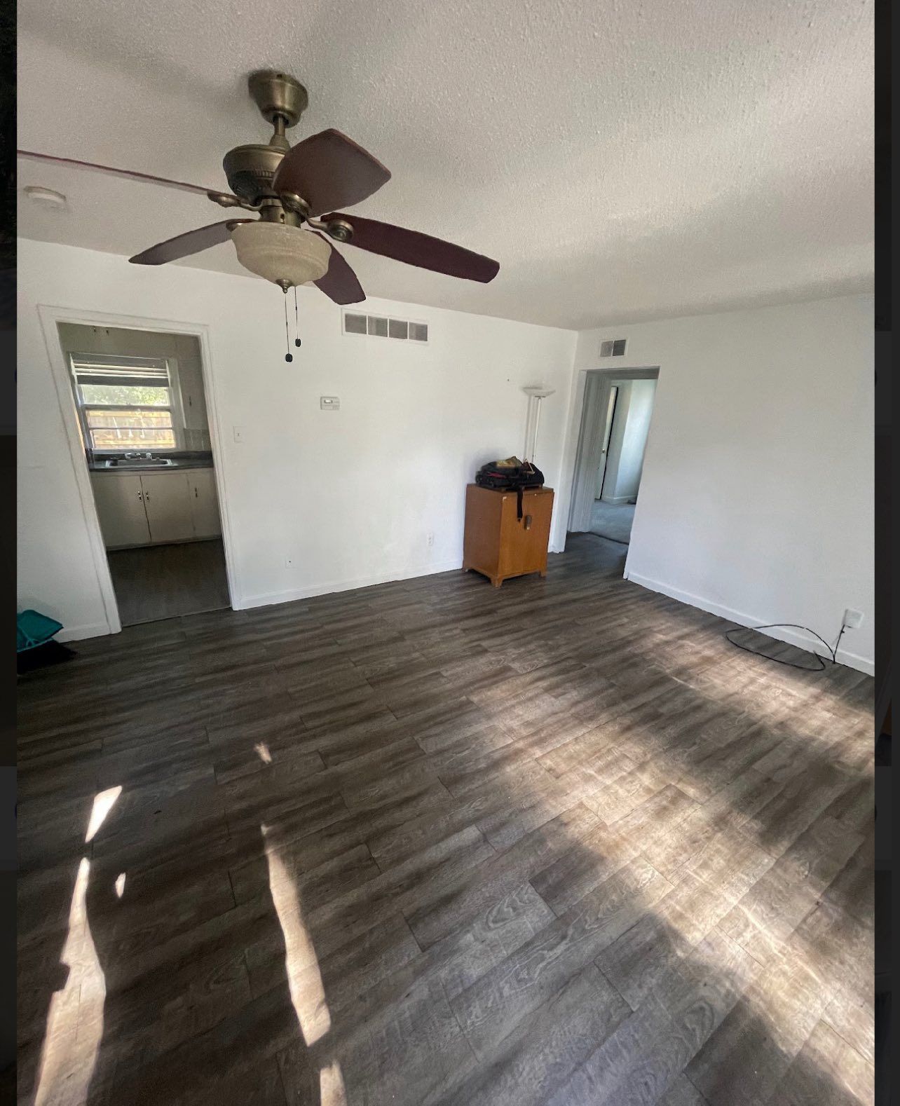 Empty living room with wood-look flooring, white walls, ceiling fan, and access to kitchen.