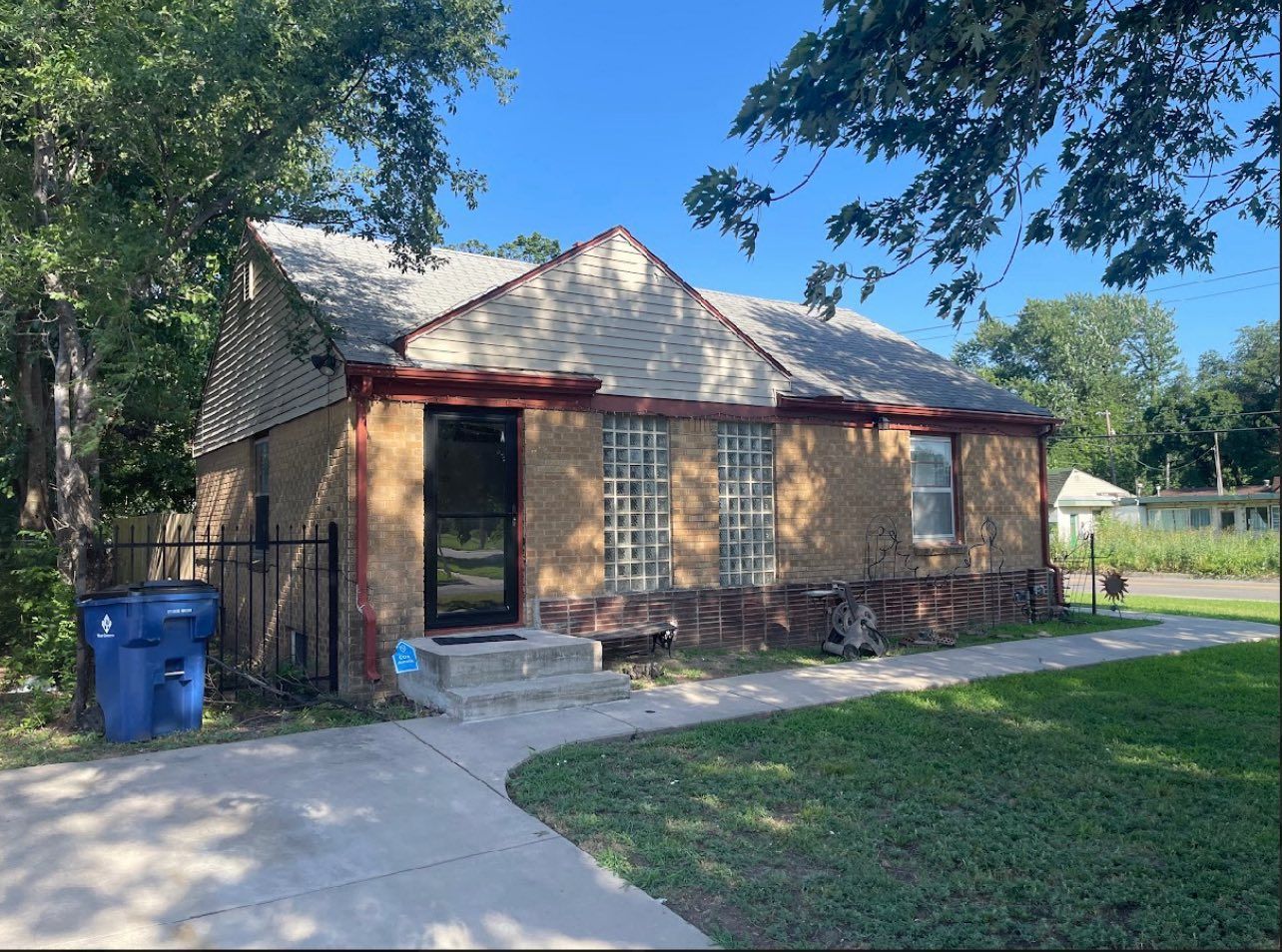 Tan brick house with a dark door, gray roof, and a blue trash can on the side.