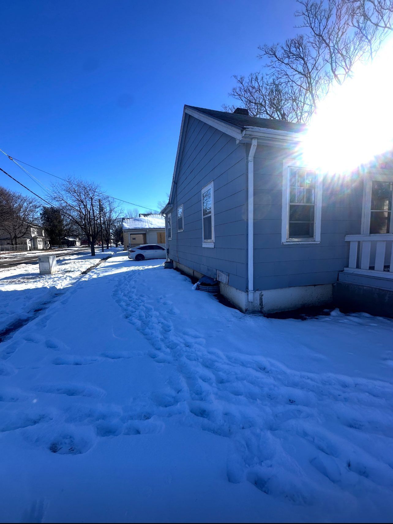 Snow-covered side of a blue house on a sunny day. Footprints in the snow lead towards the house.