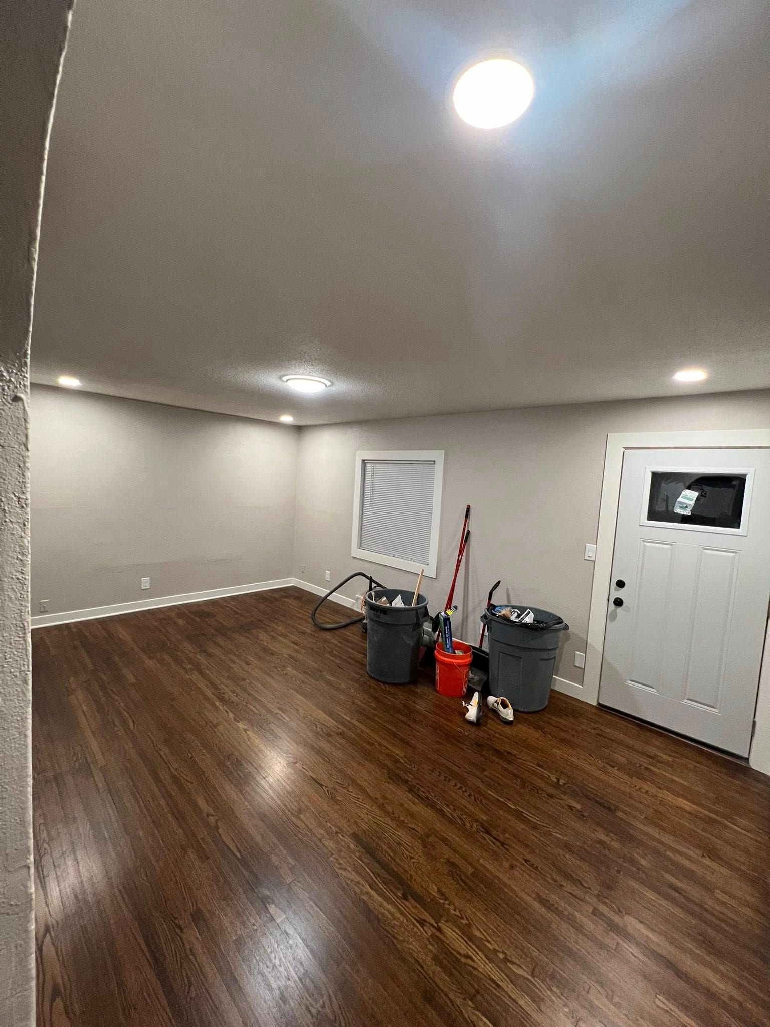 Empty room with dark wood floors, light gray walls, and a white door. Two trash cans and a mop are present.