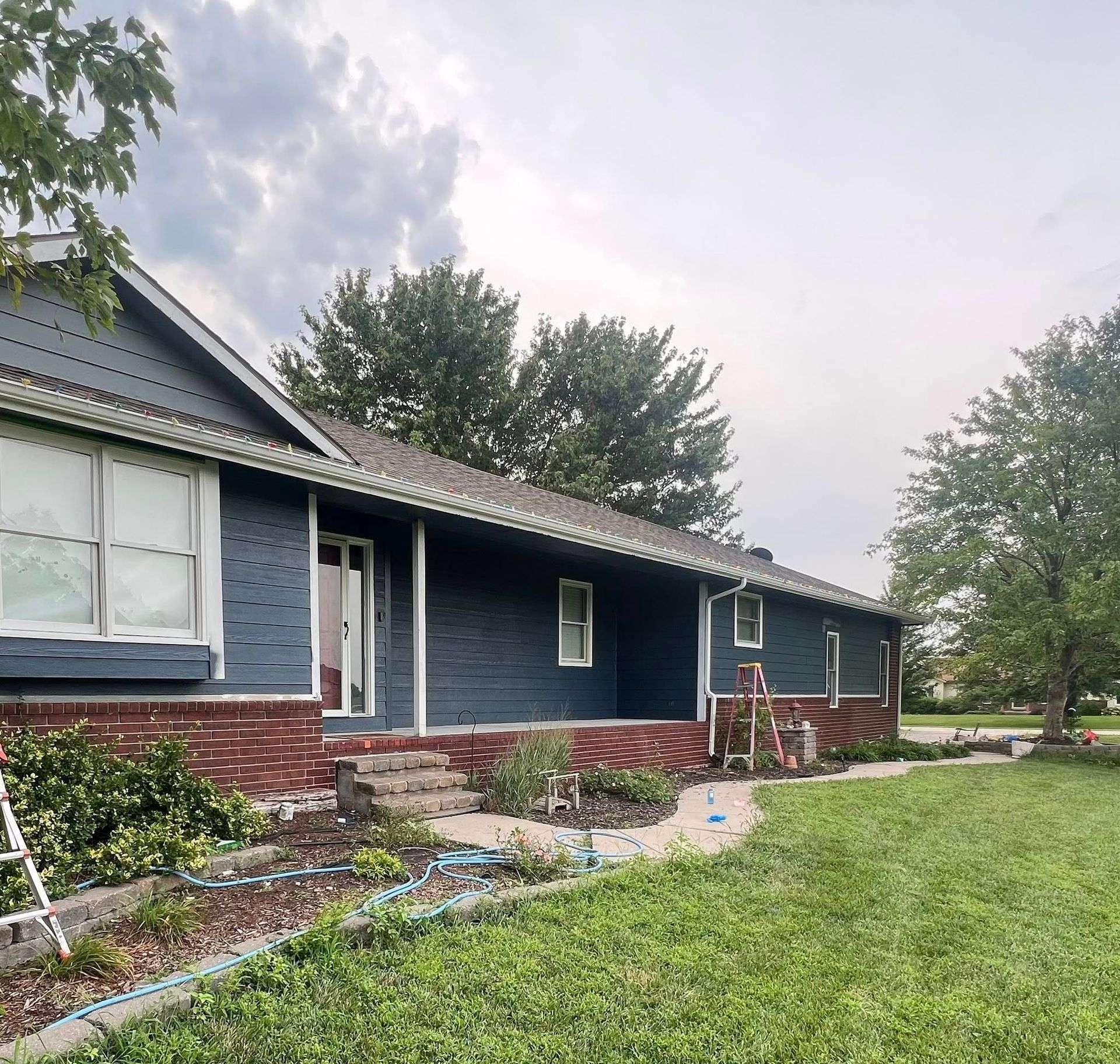 House exterior painted dark blue with brick base, under cloudy sky. Garden bed and green lawn in front.