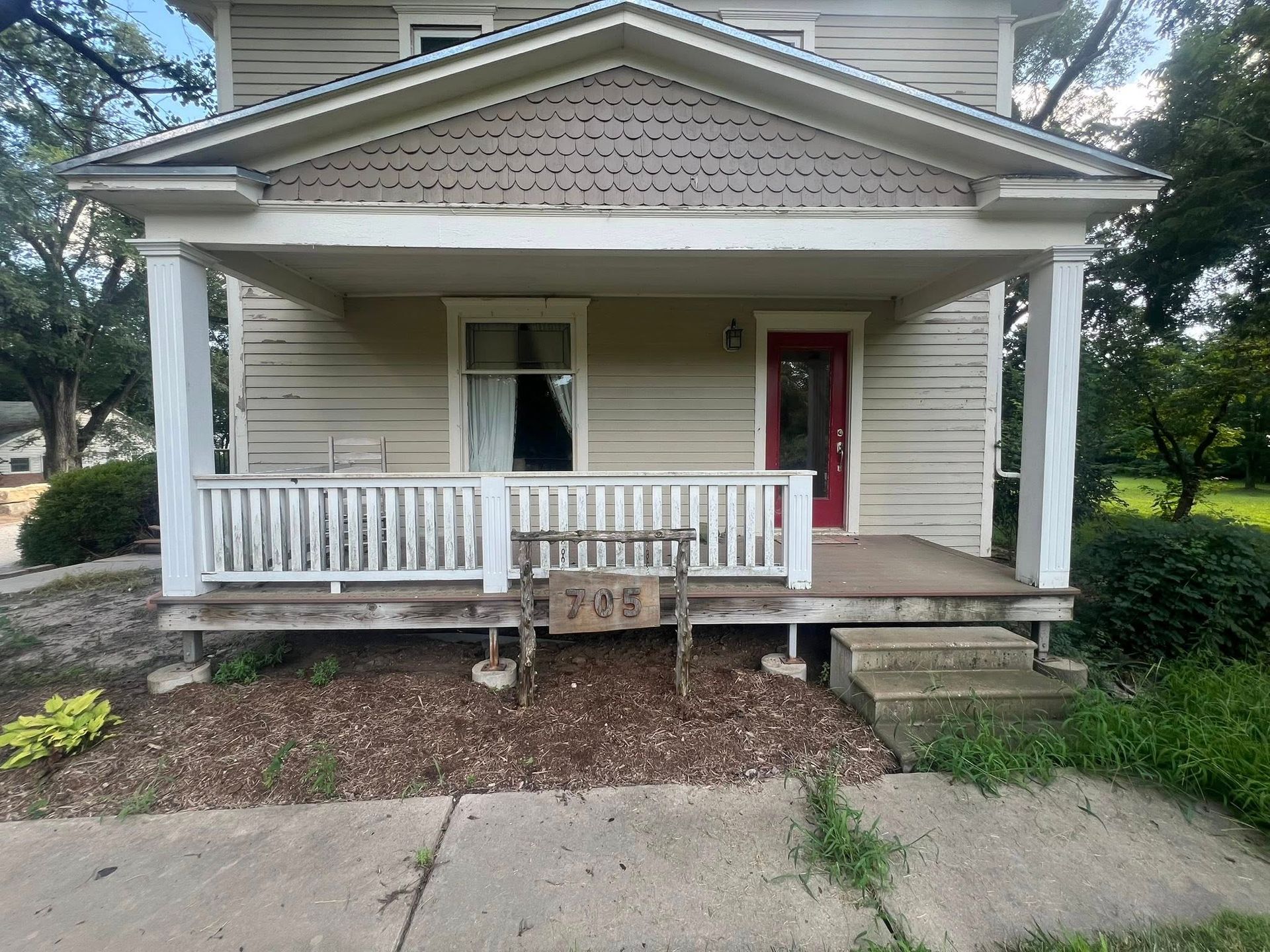A two-story house with a porch. The house has a beige exterior with a red door and white trim.