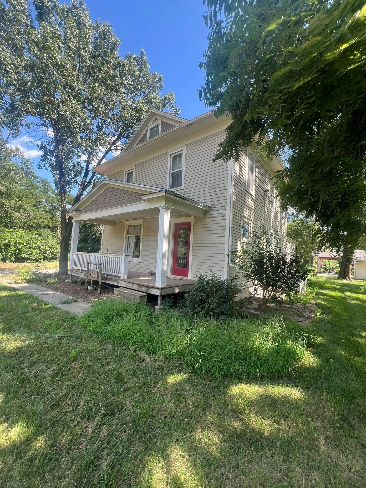 Two-story house with porch, white siding, red door, and overgrown yard on a sunny day.